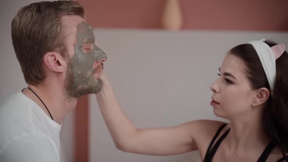 Woman Applying Clay Mask on Boyfriend Guy Face. Skin Care Routine. Couple Family Time. Cleansing