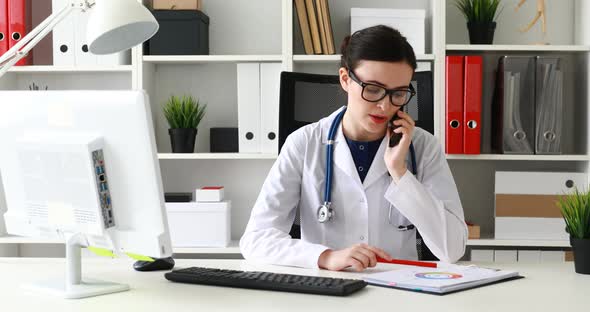 Doctor in Glasses Calls on Phone at the Workplace