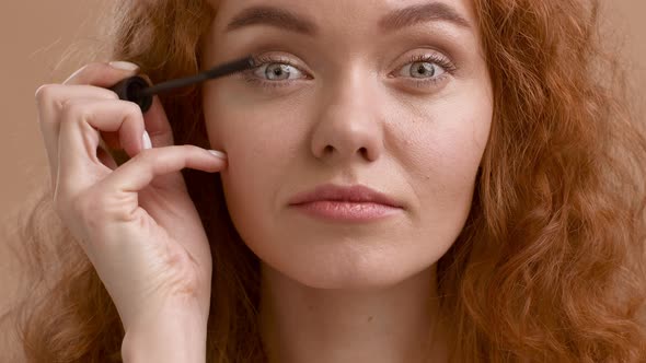 RedHaired Young Lady Applying Mascara On Eyelashes On Beige Background alt