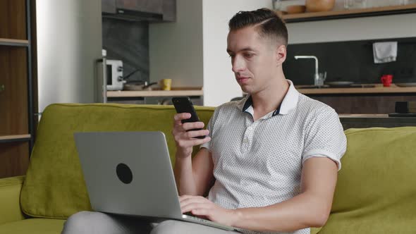 Young Busy Male Entrepreneur Uses Smartphone While Working at a Laptop Sitting at Home on the Couch alt
