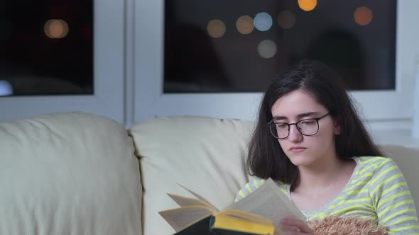 Girl with Glasses Sits on the Couch in the Evening and Enjoys Reading a Book