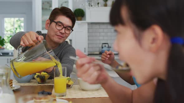Happy asian father in kitchen having breakfast pouring juice for smiling son and daughter alt
