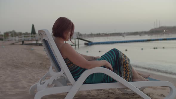 Woman in Dress Sitting on Beach Chair on the Sea Shore Watching Sunrise Over the Sea alt
