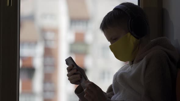 The Boy in a Protective Mask Sits on a Windowsill with the Phone in His Hands. Karatnin