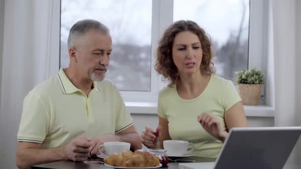 A Married Elderly Couple Discuss the News Looking Into a Laptop Over Breakfast