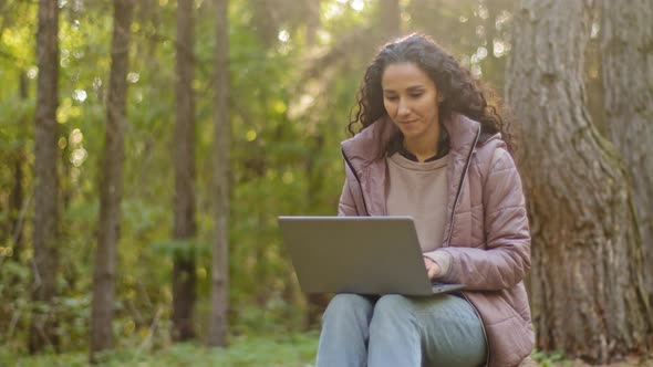 Beautiful Young Hispanic Woman with Long Curly Hair Sitting Outdoors in Autumn Forest Using Laptop alt