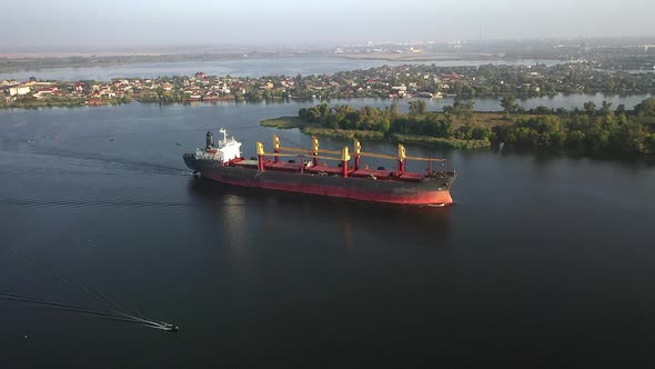 Slow Motion Areal View of Cargo Bulker Vessel Arriving Port Sailing Past Trees and Small Buildings alt