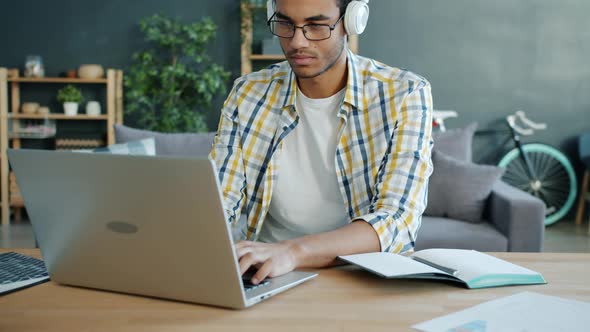 Afro-American Student Wearing Headphones Working with Laptop Typing at Home alt