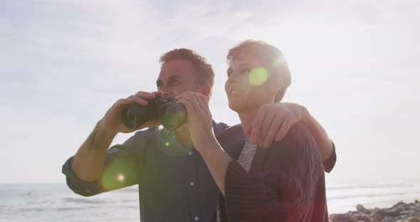 Caucasian couple enjoying free time by sea on sunny day holding binoculars alt