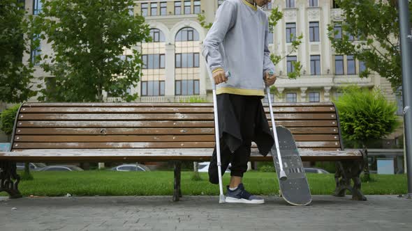 Attractive Man Sits on the Bench in the Park Putting His Crutches and Skateboard Nearby. Active Life alt