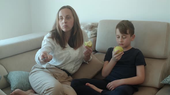 Mother and her son watching tv and eating green apples at home and sitting on sofa alt