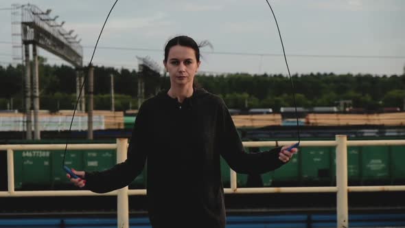 young woman on roof jumps with rope on city street against background of trains alt