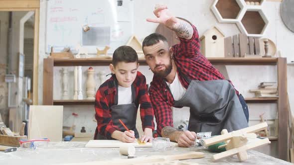 Carpenter Pointing at Something and Little Son Looks at It at Workshop