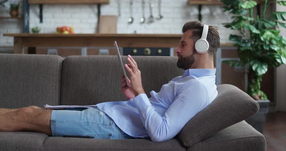Handsome Businessman in Headphones Lying in Cozy Sofa Working on Digital Tablet Looking Document alt