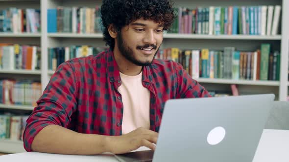 Satisfied Clever Arabian or Indian Student Guy Freelancer Sits in the Library at a Desk with Laptop alt