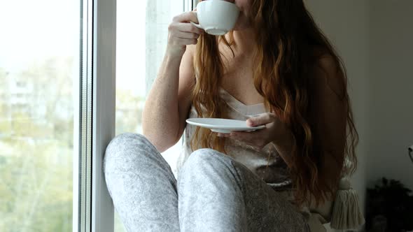 Portrait of woman drinking coffee, sitting on windowsill at window in morning