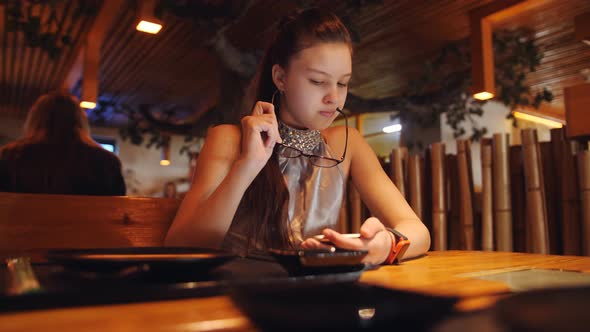 The Girl in the Restaurant Wears Glasses and Reads Messages in Social Networks on Smartphone