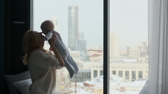 happy mom picks up and hugs baby sitting by the window