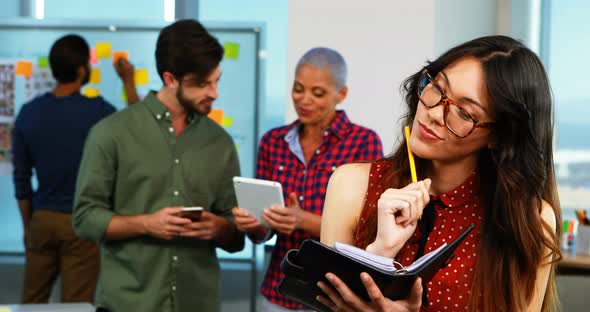 Female executive writing in organizer and colleagues discussing in background alt