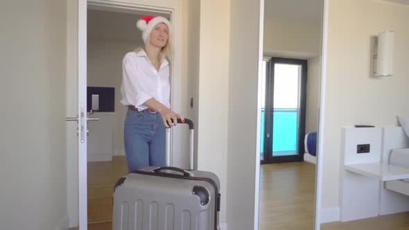 woman in a Santa Claus hat enters the hotel with a suitcase and goes to window. alt