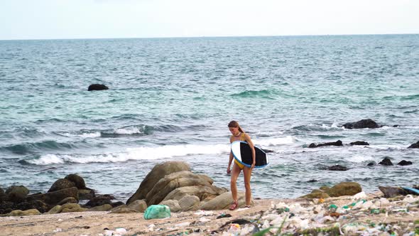 Young surfer in bikini walk in sandal on beach with trash plastic waste and ocean pollution, female alt