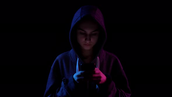 A Young Woman in a Hood with a Phone in Her Hands. Hacker Makes a Hack Through the Phone. Blue and alt