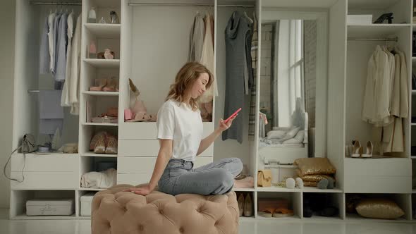 the Girl Sits on an Ottoman in the Dressing Room and Looks at the Phone