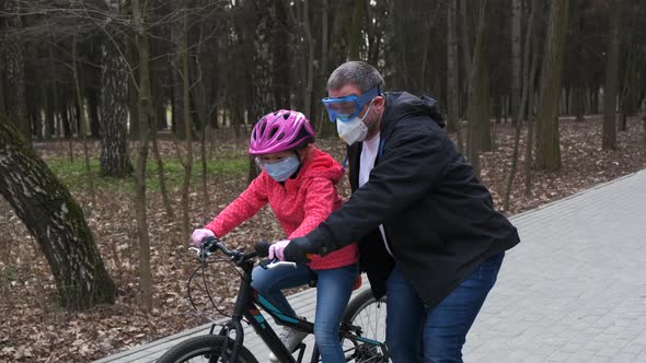 A father teaches his daughter to ride a Bicycle in a city Park. they are wearing protective helmets
