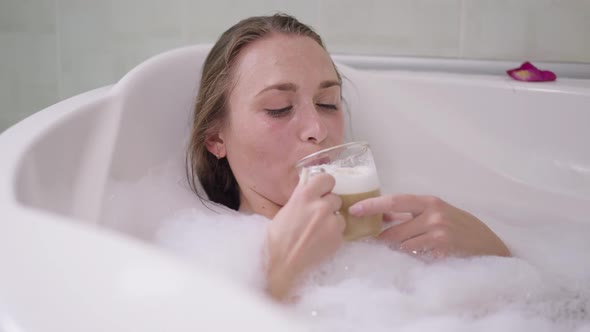 Happy Young Slim Woman Drinking Cappuccino Lying in Bath with Foam at Home alt