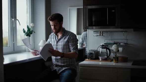 Serious Guy Reads a Paper Documents in the Kitchen By the Window alt