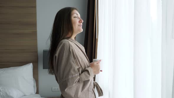Girl in Bathrobe Drinks Coffee Near Window in Hotel Room alt