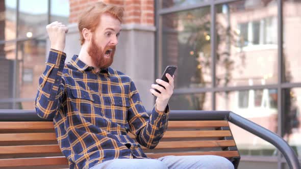 Redhead Beard Young Man Celebrating Win on Smartphone Sitting Outdoor on Bench alt