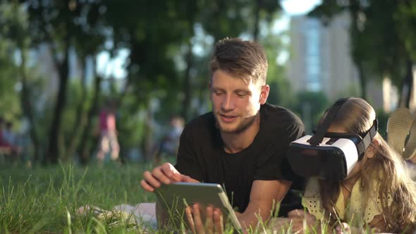 Portrait of Young Man with Tablet and Little Girl in VR Headset on the Right Lying on Green Meadow alt