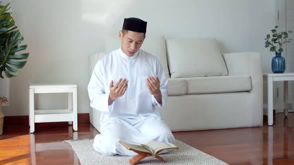 Young Muslim man wearing Islamic clothes reading Quran and praying alt