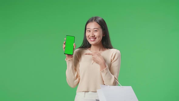 Woman With Shopping Bags Holding And Pointing Green Screen Mobile Phone In Front Of Green Screen alt