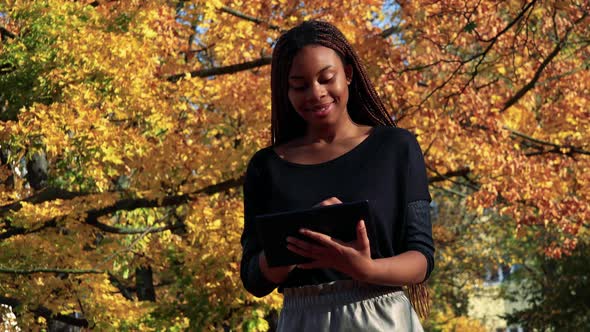 Young Beautiful Black Woman Works on the Tablet in Autumn Park alt