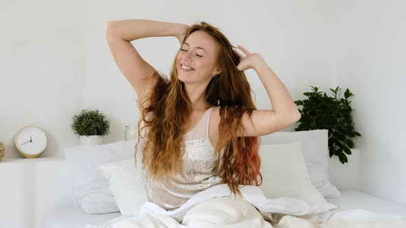 Happy woman in pajamas sits on bed and stretches with pleasure after sleeping
