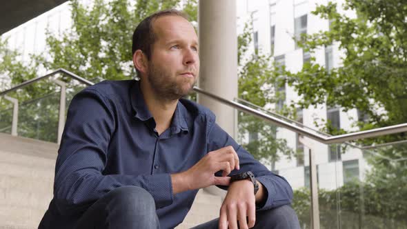 A Caucasian Man Works on a Smartwatch As He Sits on a Staircase in an Urban Area