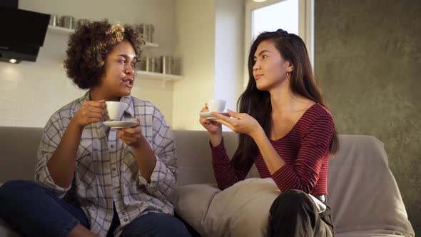 Two AfricanAmerican and Asian Girlfriends Laugh While Having Tea Together at Home alt