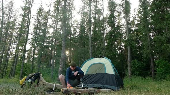 Disabled man with prosthetic limb is sitting on in forest near fire and rest. alt