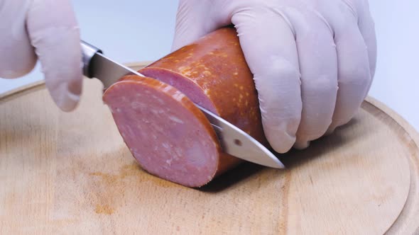 The cook cuts meat sausage into large pieces for a sandwich on a wooden table. A thick cured salami