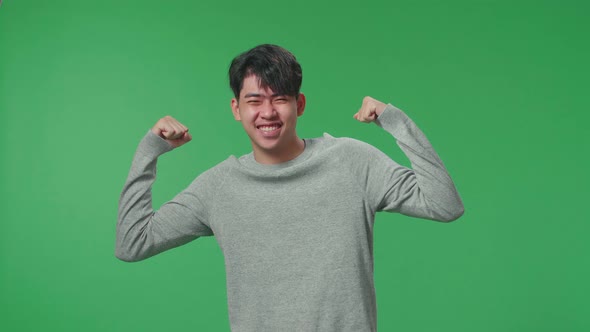 A Smiling Asian Man Flex Muscle While Standing In Front Of Green Screen Background alt