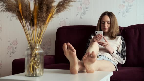 Barefoot Girl Sits On Couch And Holds Her Legs On Table, She Is Reading News On Mobile Phone