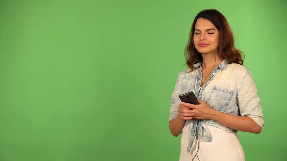 A Young Caucasian Woman Listens to Music on a Smartphone with Earphones on and a Smile Green Screen