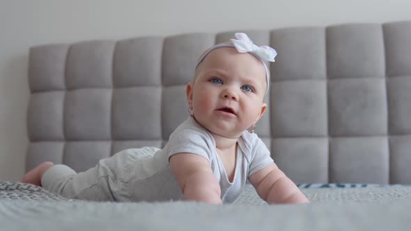 Baby Girl in Gray Clothes with a Cute Bow on His Head Lying on the Bed Tummy Down and with a Serious alt