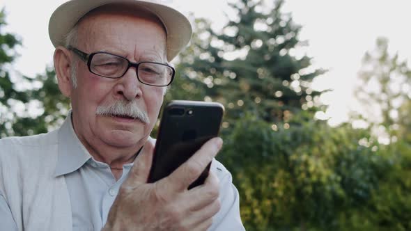 Portrait of Senior Man with Glasses in Hat Chats on Phone's Web Camera in Park alt