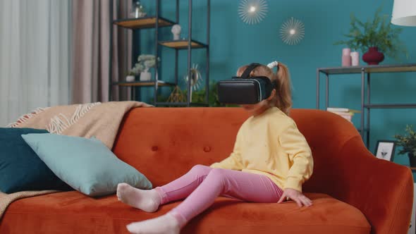 Toddler Girl Sitting on Home Sofa Using Virtual Reality Headset Helmet App to Play Video 3D Game alt