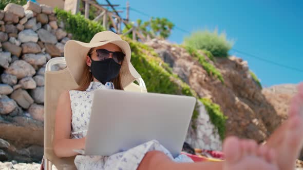 Lady in Face Mask Works Online on Laptop Relaxing on Beach in Summer in Greece