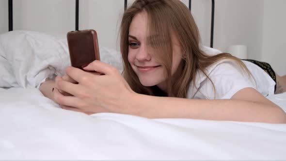 Young Happy Smiling Woman Using Her Phone While Lying in Bed in the Morning