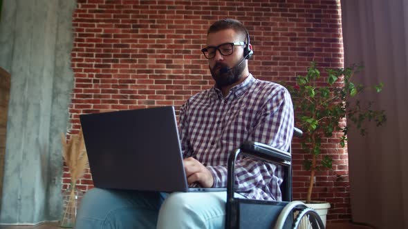 Man in Glasses is Sitting on Wheelchair and Working on Laptop with Headset alt